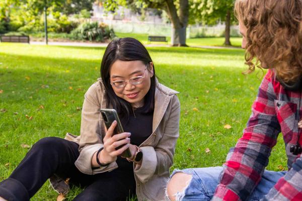 A young person sitting on the grass in a park. She is looking at her phone.