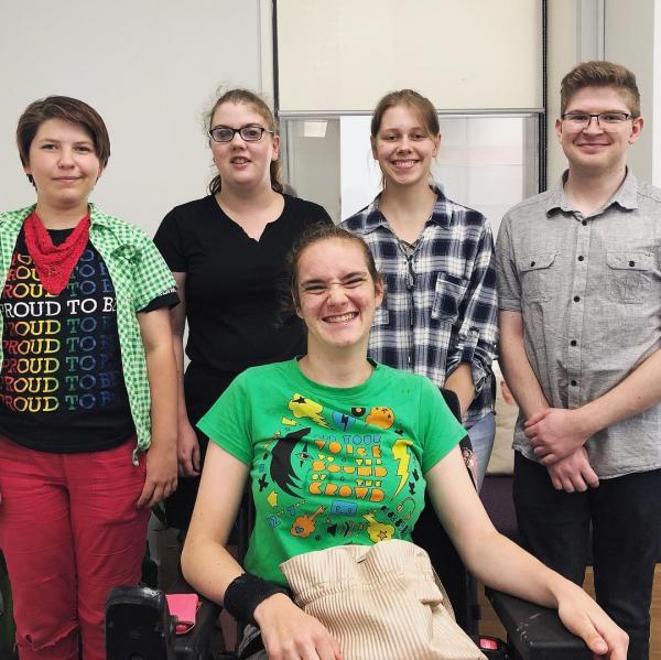 Five young people smile together indoors, with one person in a wheelchair at the front and the others standing behind, showing a sense of friendship and inclusion.