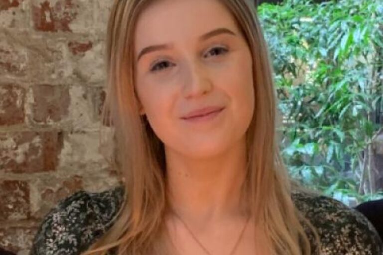 White person with blonde hair wearing a necklace and smiling